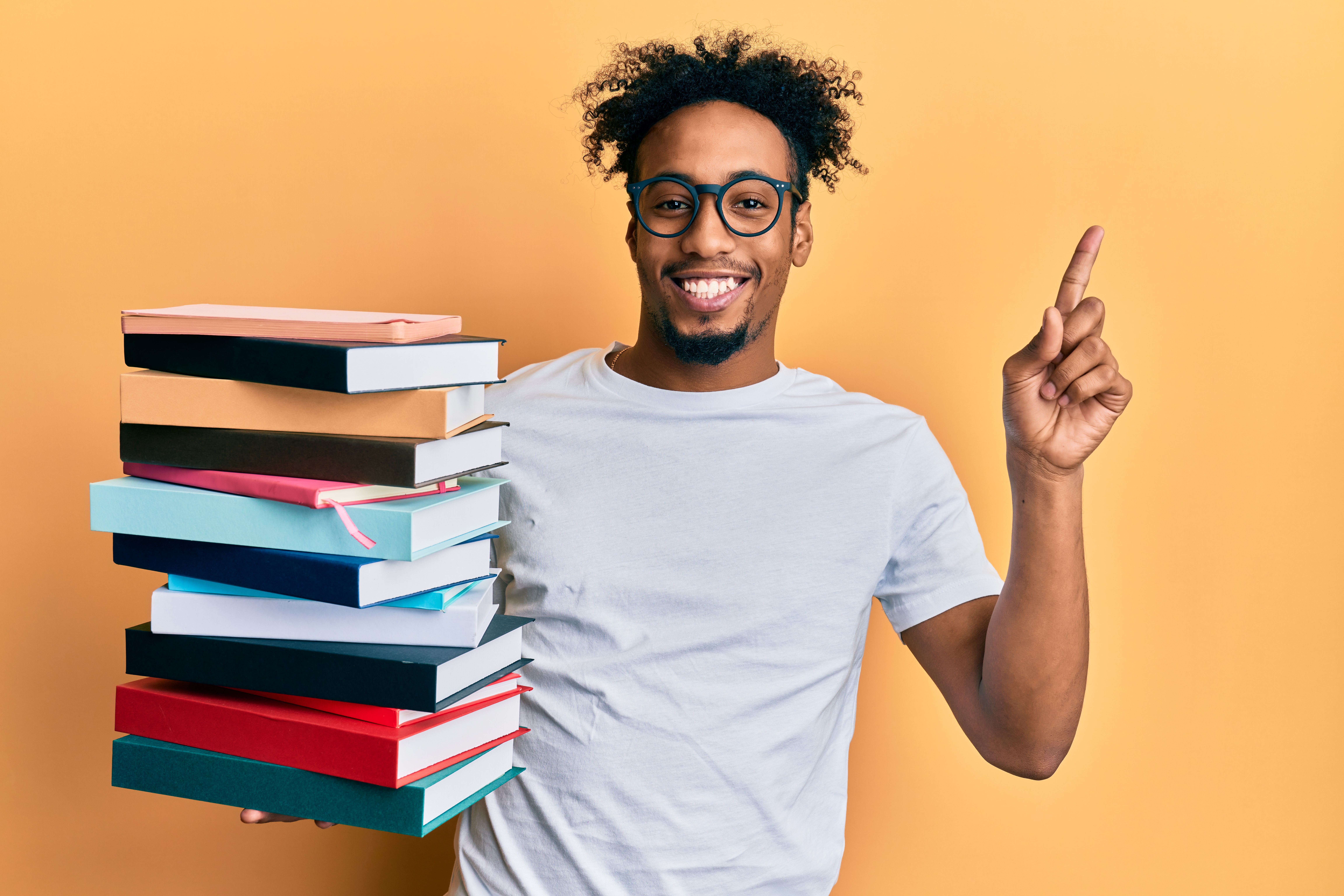 young african american man with beard holding a pile of books smiling happy pointing with hand and finger to the side